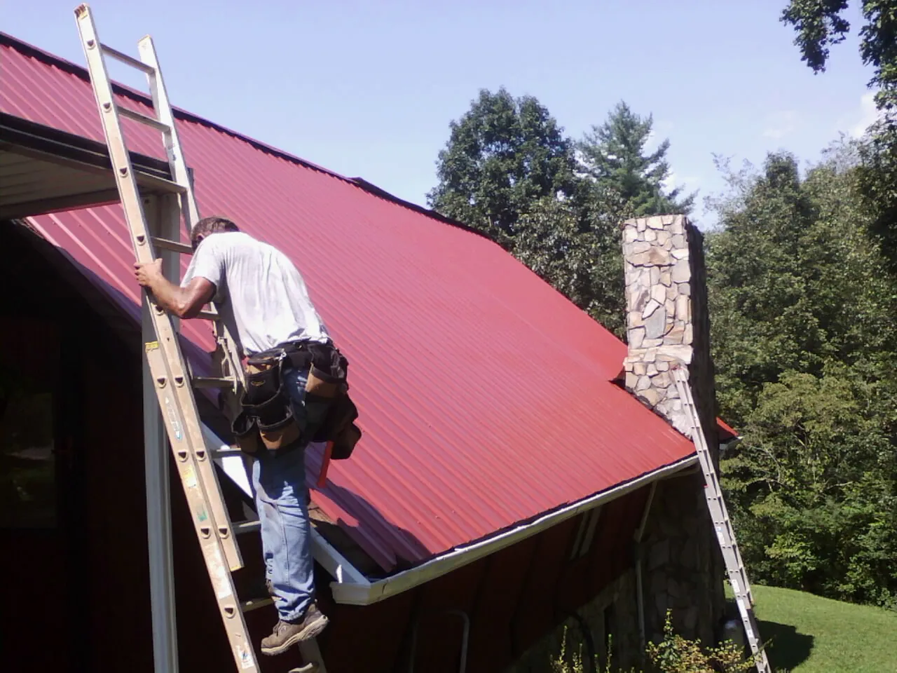 Roofer on a ladder beside a red metal roof and stone chimney — Soffit & Fascia Repair by Roofing Brothers in Navarre